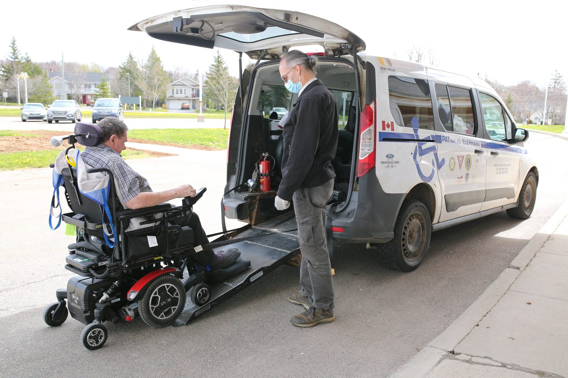 Pat & The Elephant accessible wheelchair van with ramp deployed, driver assisting a passenger in a wheelchair into the vehicle on a sunny day